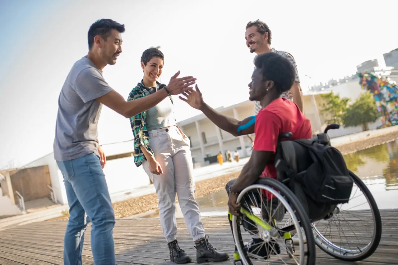 A young man in wheelchair, having a handshake with three of his white friends.