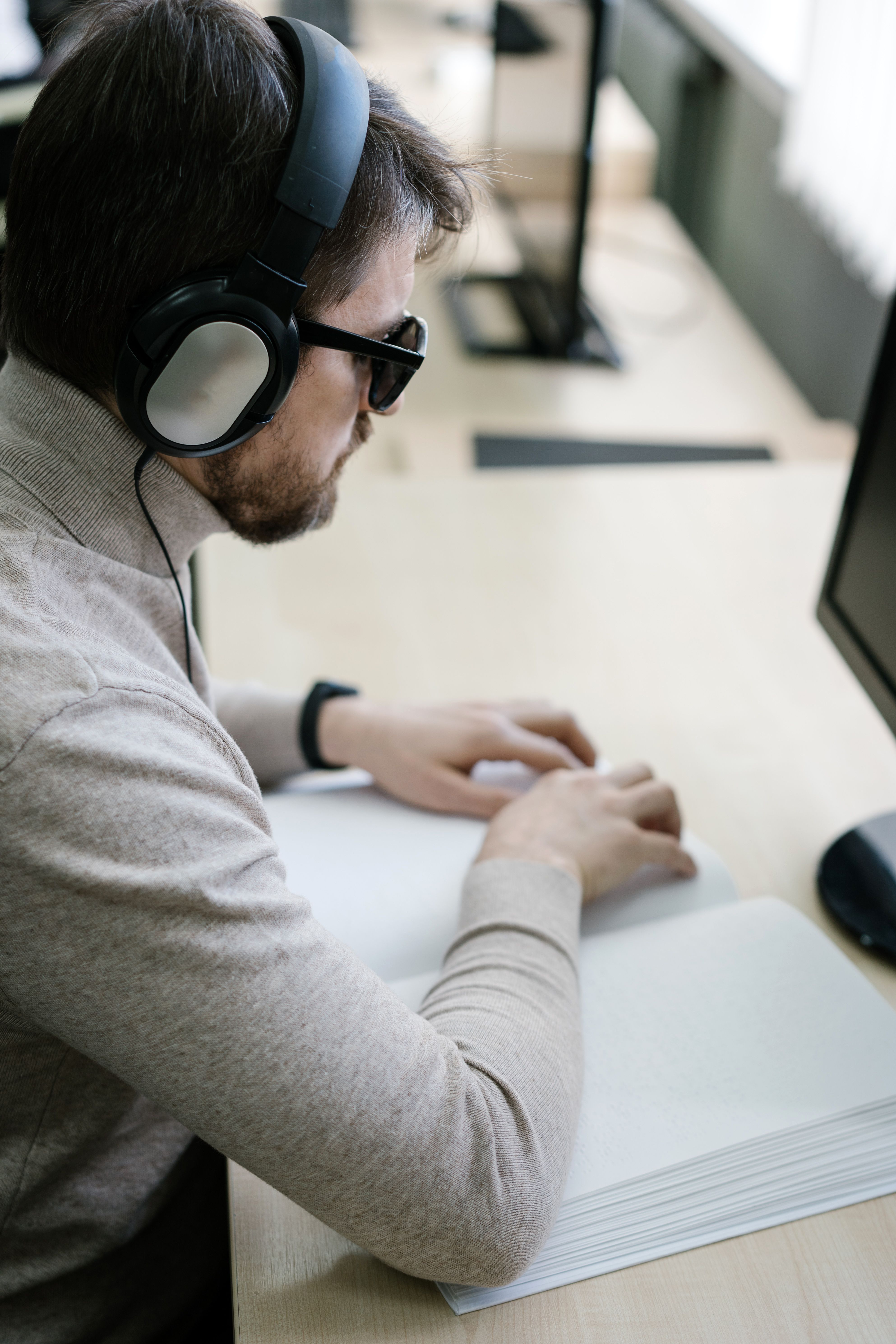 Blind employee wearing headphones and using Braille materials at a computer workstation.