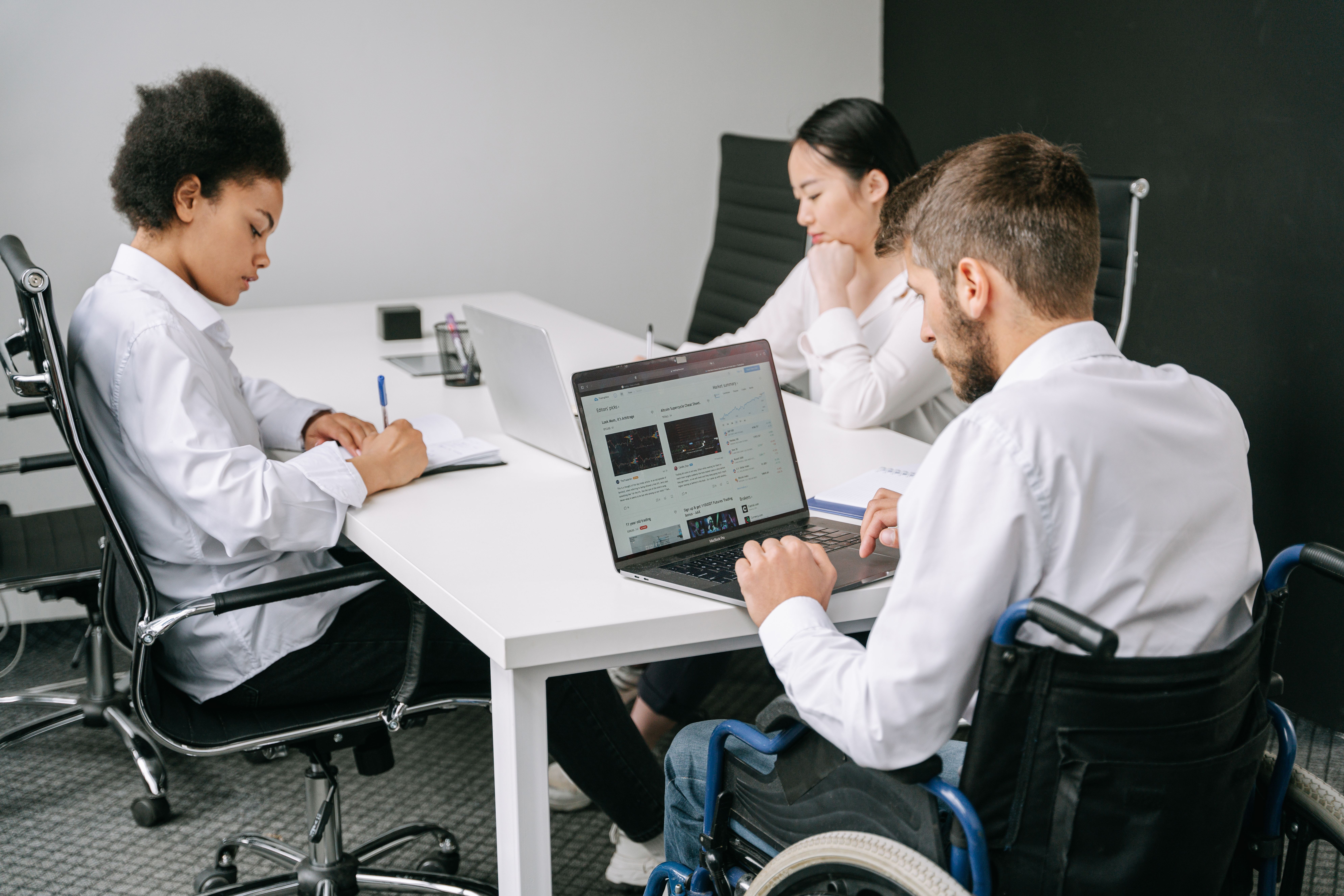Three colleagues, including two wheelchair users, working together around a table with laptops and notes.