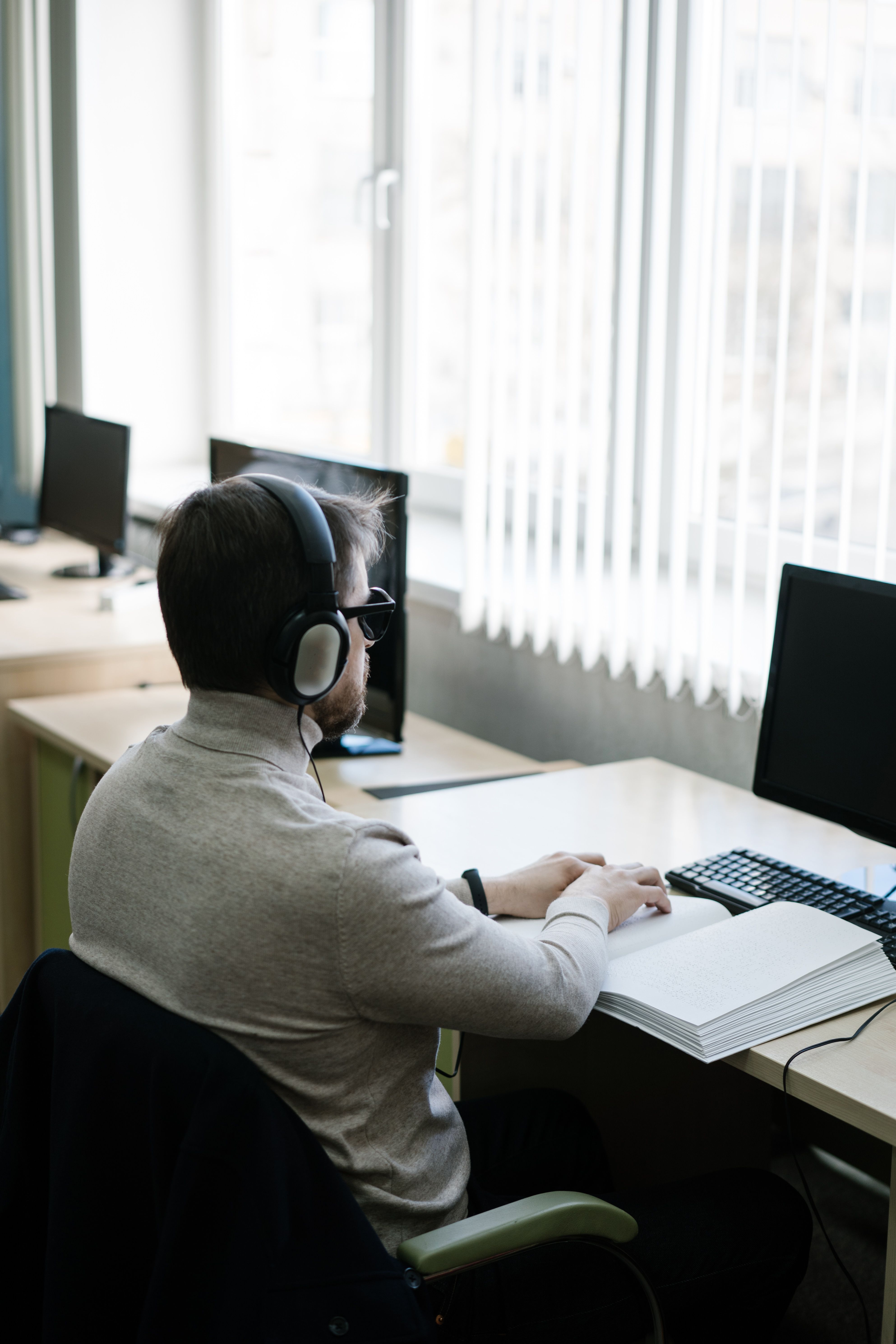 Blind employee wearing headphones and working at a computer beside Braille materials in an office.