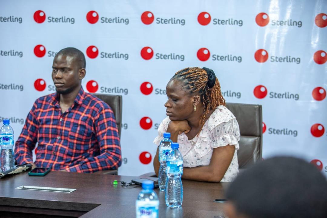 Two blind beneficiaries, a man and a woman, sit at a conference table during a Sterling Bank event, with Sterling branding visible on the backdrop and water bottles on the table.